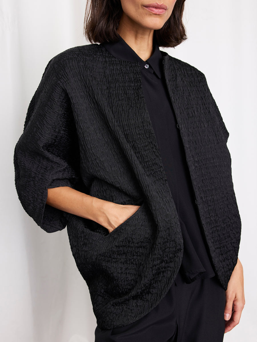 A woman models the Zero + Maria Cornejo Rio Shrug in black, layered over a matching collared shirt and pants, standing with one hand in her pocket against a plain white background.