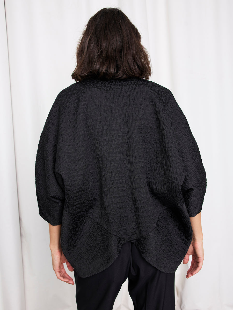 A person with shoulder-length dark hair, seen from behind, wears the Rio Shrug by Zero + Maria Cornejo with black pants, standing in front of a white curtain.