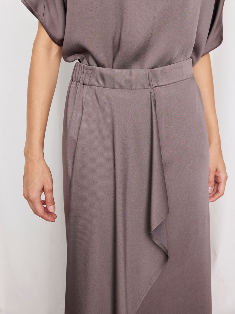 A person models the Zero + Maria Cornejo One Skirt—a loose, taupe dress with short sleeves and an asymmetrical ruffle skirt—against a plain light background.