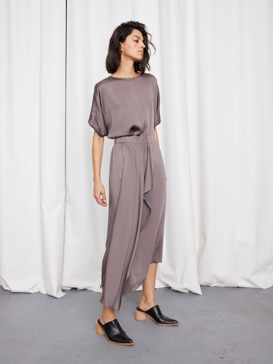 A woman stands before white curtains wearing the “One Skirt” by Zero + Maria Cornejo, a loose taupe asymmetrical ruffle skirt dress with short sleeves, paired with black mules. She gazes to the side in a relaxed pose.