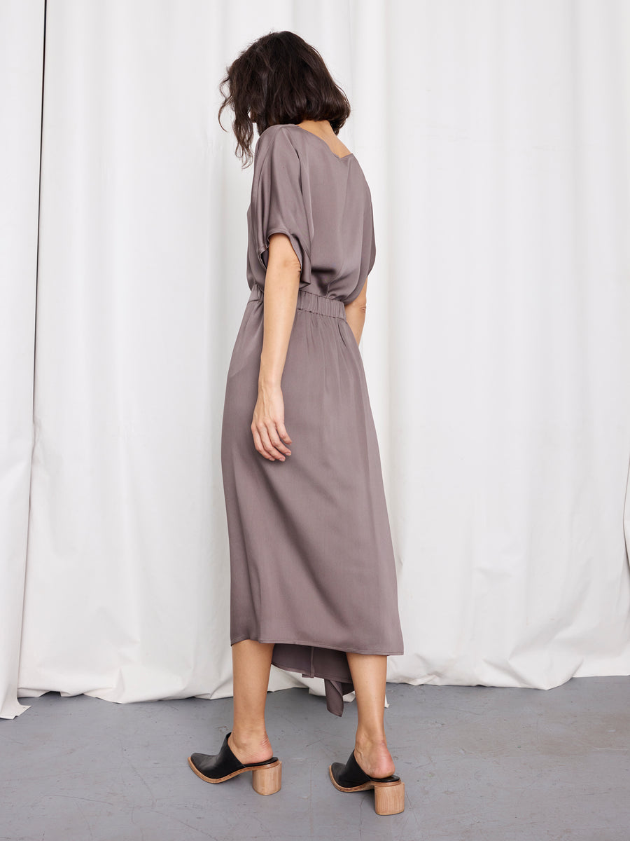 A woman faces away, wearing Zero + Maria Cornejo’s One Skirt—a loose, taupe mid-length piece with short sleeves—paired with black heeled mules. She stands before white curtains on a gray floor.