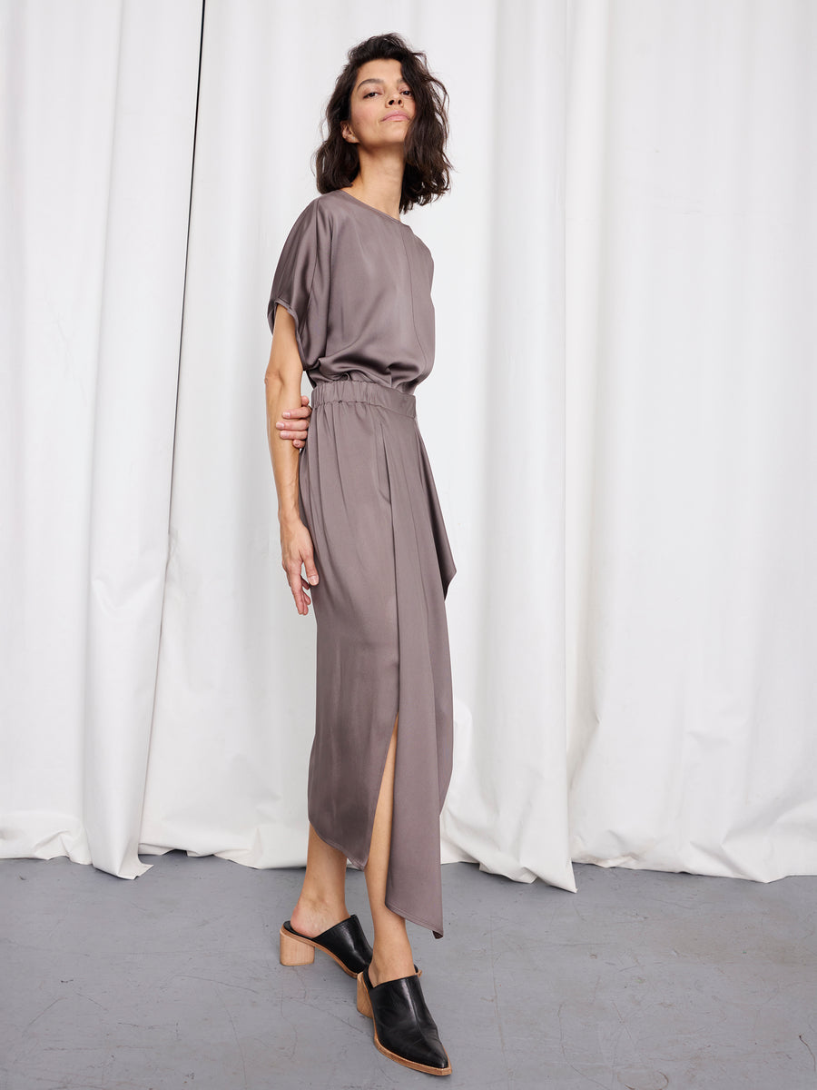 A woman with short dark hair stands confidently in front of white curtains, wearing Zero + Maria Cornejo's One Skirt—a loose, short-sleeved taupe dress with an elastic waist and side slit—paired with black heeled mules.