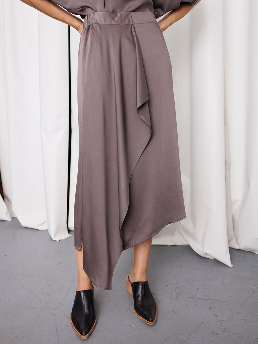 A person wearing the Zero + Maria Cornejo One Skirt in taupe and black pointed-toe block heels stands on a gray floor, with white curtains in the background.
