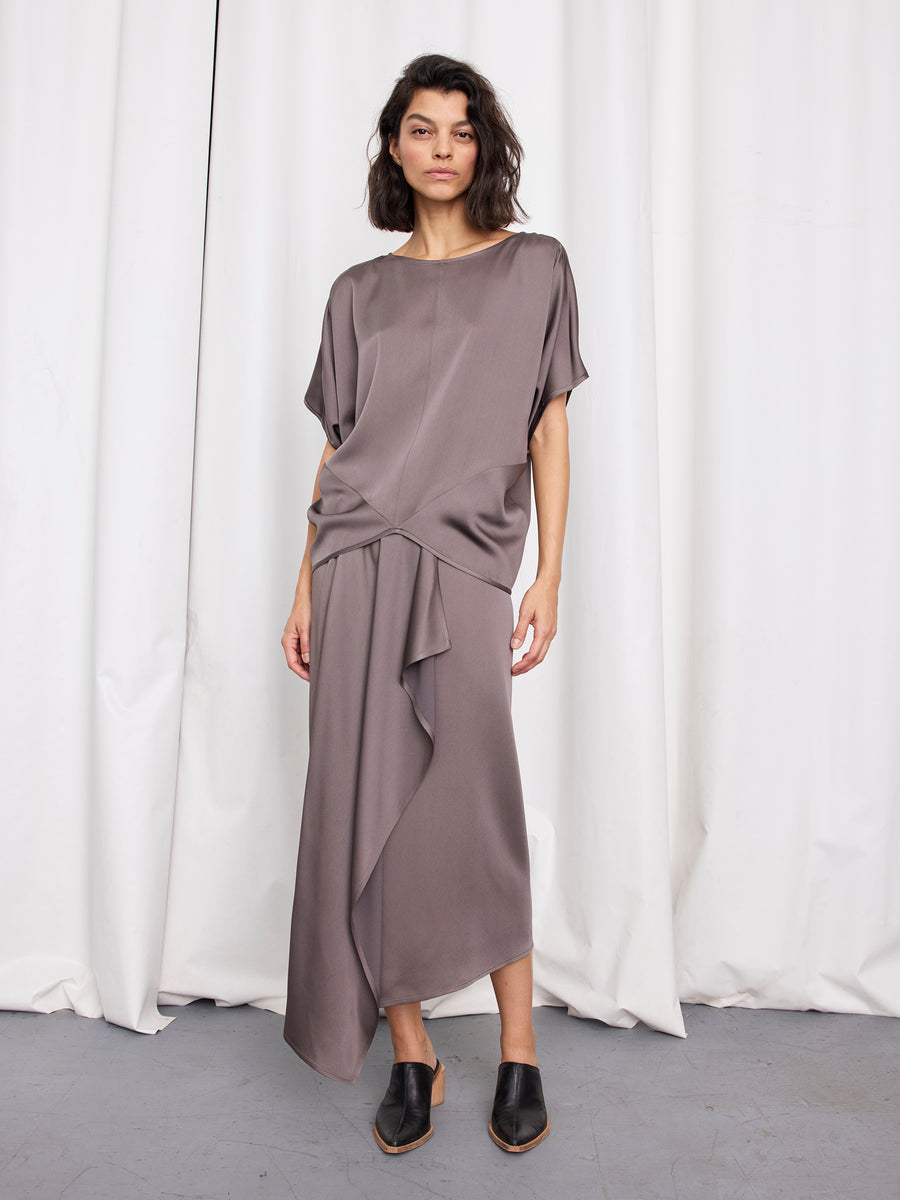 A woman with wavy dark hair stands before white curtains, wearing the taupe Rio Top by Zero + Maria Cornejo, an asymmetrical woven draped cap-sleeve piece made in New York, paired with black mules. She gazes at the camera neutrally.