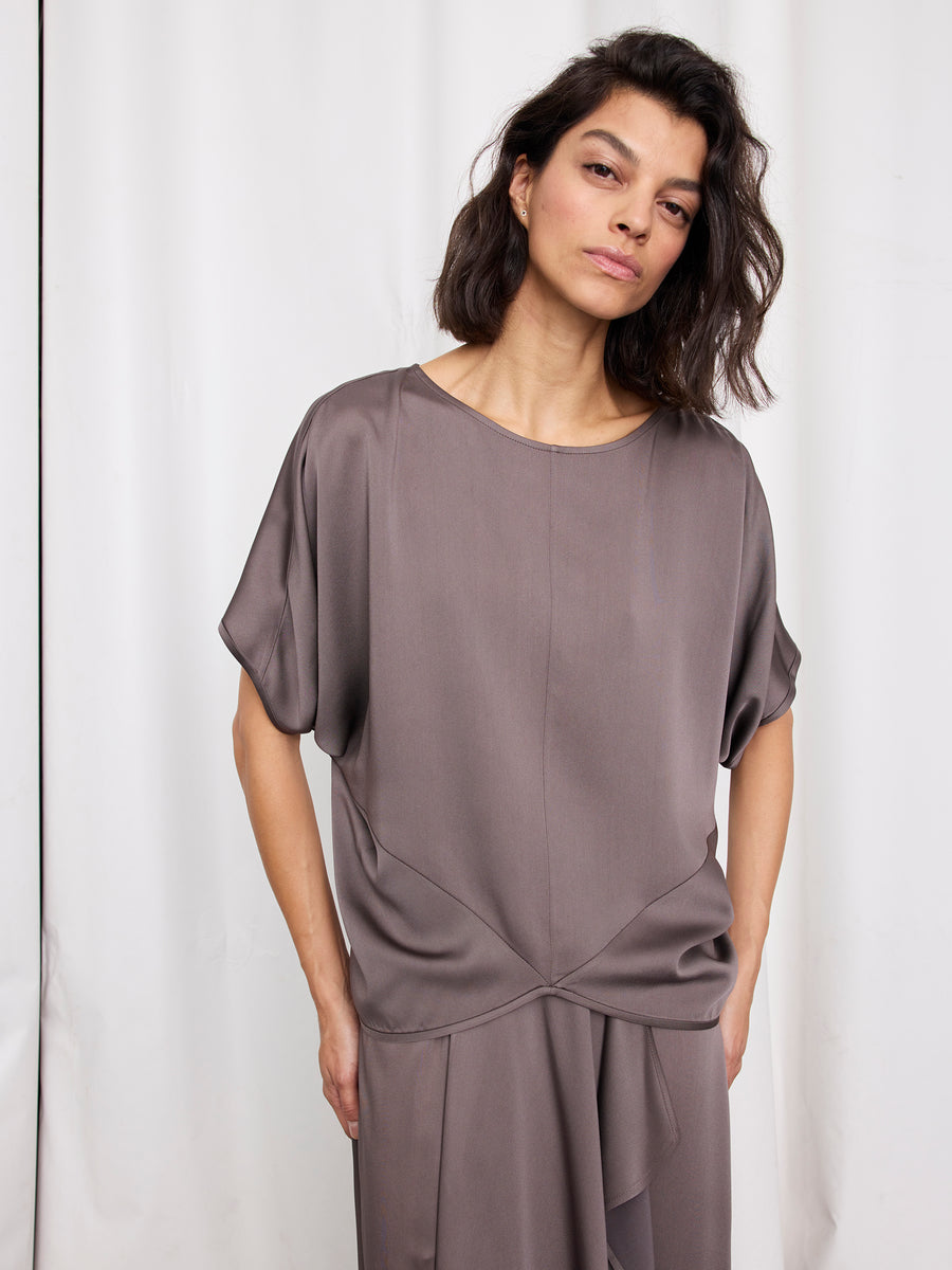 A woman with wavy dark hair wears the Rio Top by Zero + Maria Cornejo, a woven draped piece with cap sleeves, paired with a matching skirt, posing before a plain white curtain. This effortlessly chic look is made in New York.