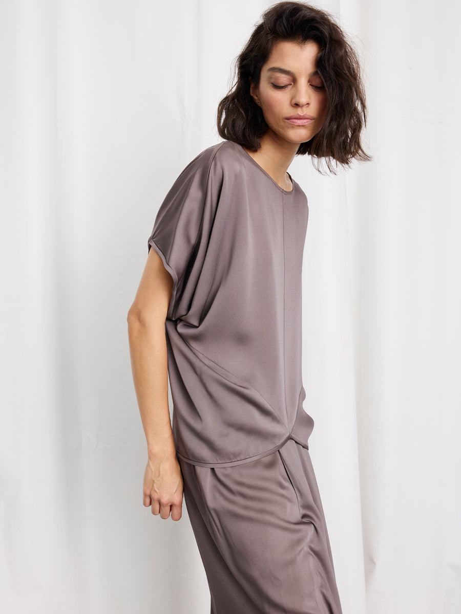 A woman with shoulder-length dark hair wears the Zero + Maria Cornejo Rio Top in taupe with matching pants, standing against a white curtain background and looking down with a neutral expression.