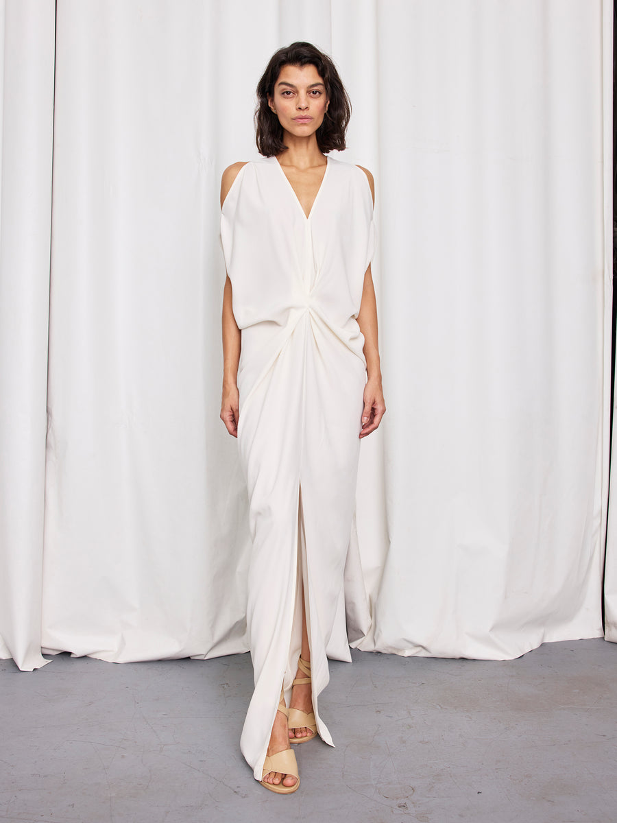 A woman wearing the Zero + Maria Cornejo Long Miu Dress—a full-length, white sleeveless piece with a knotted waist and front slit—stands before white curtains in beige open-toed heels, her dark hair framing her hourglass silhouette.