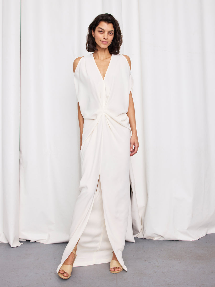 A woman with shoulder-length dark hair wears the Zero + Maria Cornejo Long Miu Dress—a sleeveless, white full-length gown with a plunging neckline and twisted knot waist—paired with beige open-toe heels, standing before white curtains.