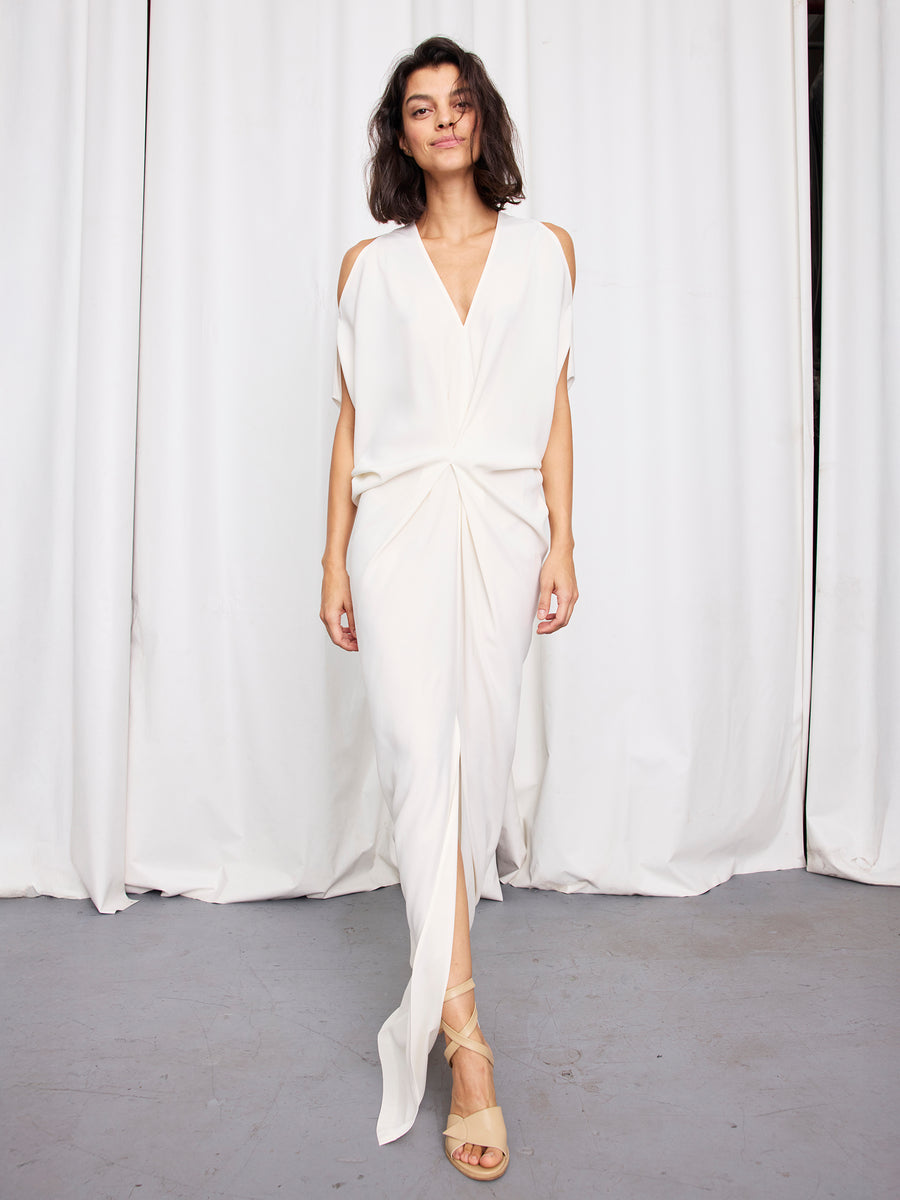 A woman with tousled dark hair smiles in Zero + Maria Cornejo’s Long Miu Dress—a full-length white dress with cut-out shoulders and a front slit—paired with strappy nude heels, standing before white curtains on a gray floor.