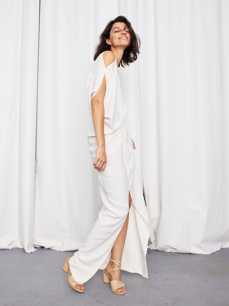 A woman with dark hair smiles in the Long Miu Dress by Zero + Maria Cornejo—an elegant, off-white full-length gown with a slit and a graceful hourglass silhouette. She pairs it with beige heeled sandals in front of white curtains.