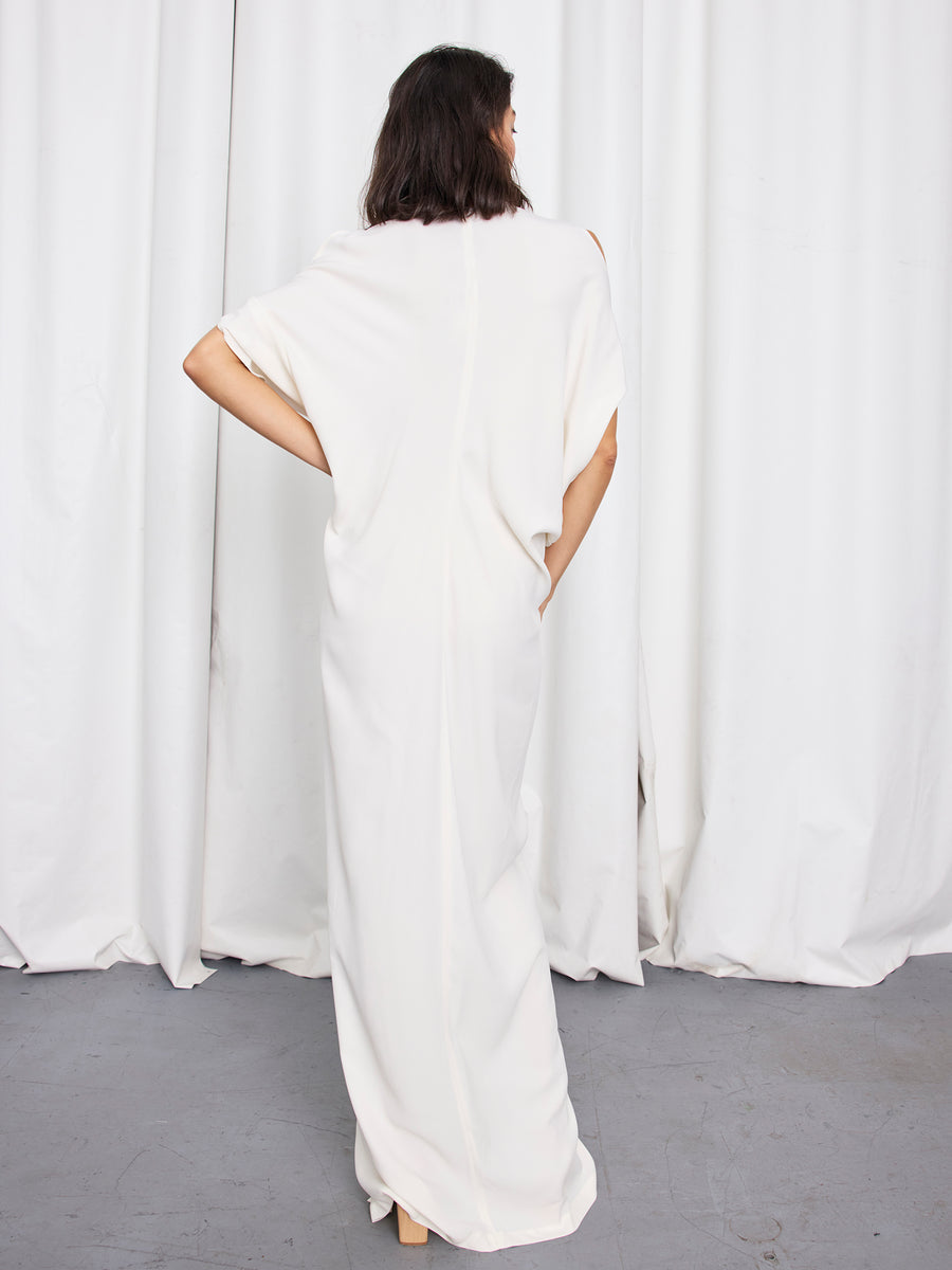 A person stands with their back to the camera, wearing the Long Miu Dress by Zero + Maria Cornejo—a loose-fitting, full-length white dress with short sleeves—against a backdrop of white curtains and a gray floor.