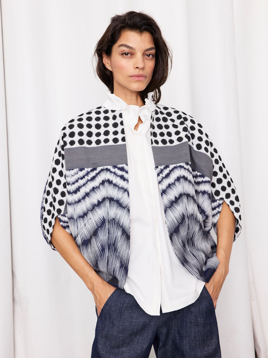 A woman with dark hair and a neutral expression stands against a white curtain, wearing a white blouse, dark pants, and the Zero + Maria Cornejo Rio Shrug—a graphic jacquard jacket with black polka dots and wavy blue lines.