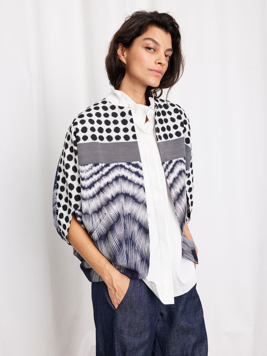 A person with medium-length dark hair stands with hands in pockets, wearing the Rio Shrug by Zero + Maria Cornejo—a graphic black and white jacquard jacket—over a white shirt and loose dark pants against a plain white background.