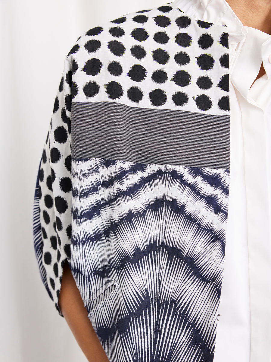A close-up of a person wearing a white collared shirt and the Rio Shrug by Zero + Maria Cornejo, a short-sleeved jacket with black dots on white, a gray horizontal stripe, and geometric black-and-white zigzag patterns.