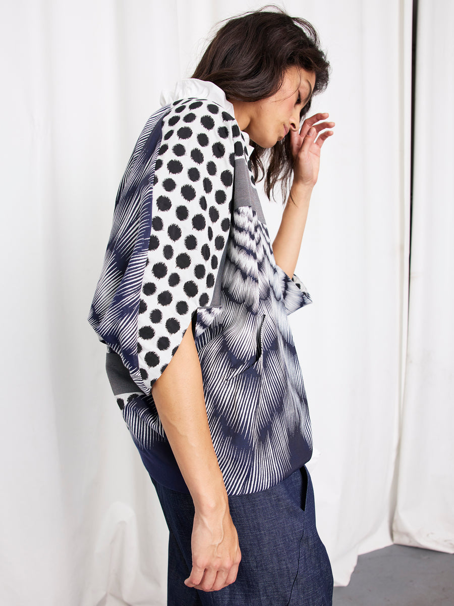 A person stands sideways against white curtains wearing the Zero + Maria Cornejo Rio Shrug, a short-sleeved outerwear with black and white polka dots and abstract designs, paired with loose blue pants. Their dark hair is loose and hand raised near their face.