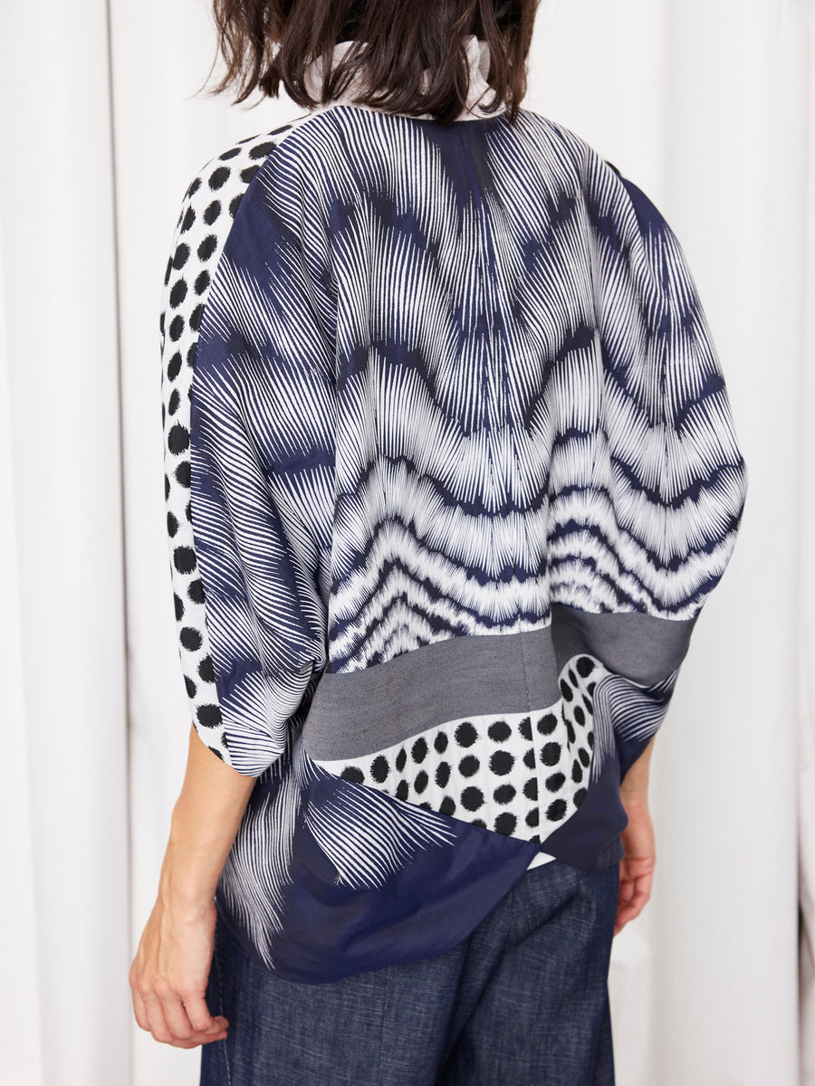 A person shown from the back wears the Zero + Maria Cornejo Rio Shrug—a loose, graphic jacquard jacket with wavy blue and white lines and black polka dots—paired with dark blue pants against a light background.