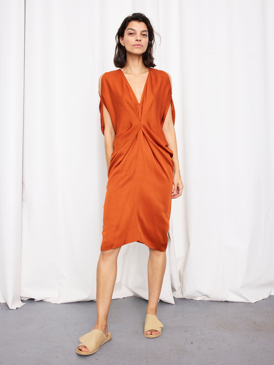 A woman with shoulder-length dark hair and a neutral expression wears the Zero + Maria Cornejo Miu Dress, a loose rust-orange knee-length style with a deep V-neckline, and beige slide sandals. She stands on a gray floor before white curtains.