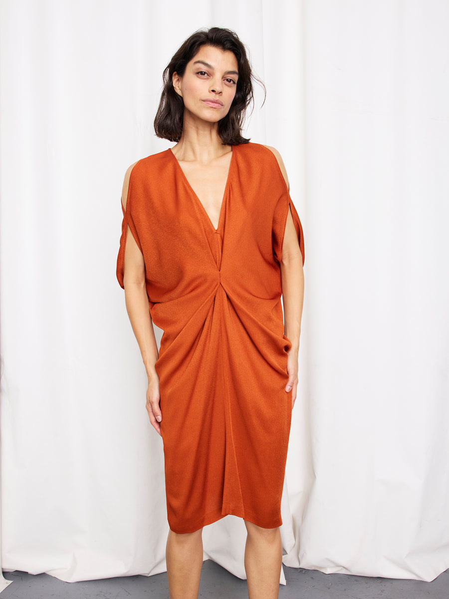 A woman with dark shoulder-length hair and a neutral expression stands against a white curtain, wearing the Zero + Maria Cornejo Miu Dress—a rust-colored, knee-length sustainable viscose dress with a deep V-neckline and gathered waist.