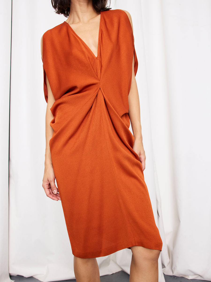 A woman wears the Zero + Maria Cornejo Miu Dress—an orange sustainable viscose, knee-length dress with a deep V-neck and gathered waist twist—standing before white curtains. Her head is out of the frame.