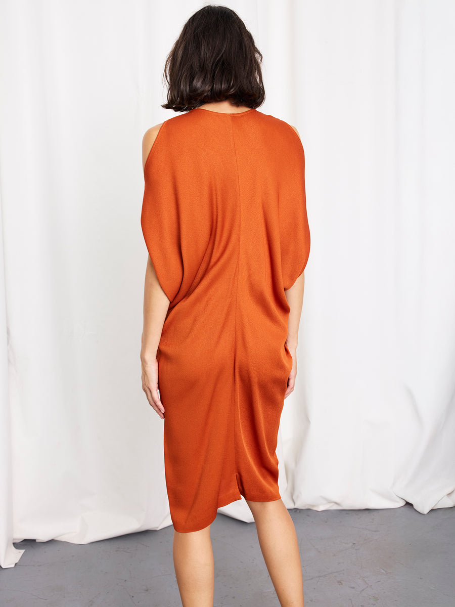 A woman with short dark hair stands facing away, wearing the Miu Dress by Zero + Maria Cornejo—an orange, knee-length sustainable viscose piece. The background features a white eco drape over a gray floor.