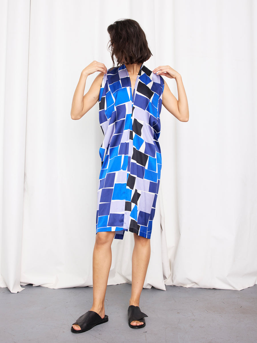 A person stands on a grey floor before white curtains, wearing the Issa Dress by Zero + Maria Cornejo—a sleeveless blue and black graphic knee-length dress—paired with black slide sandals, hands touching the collar.