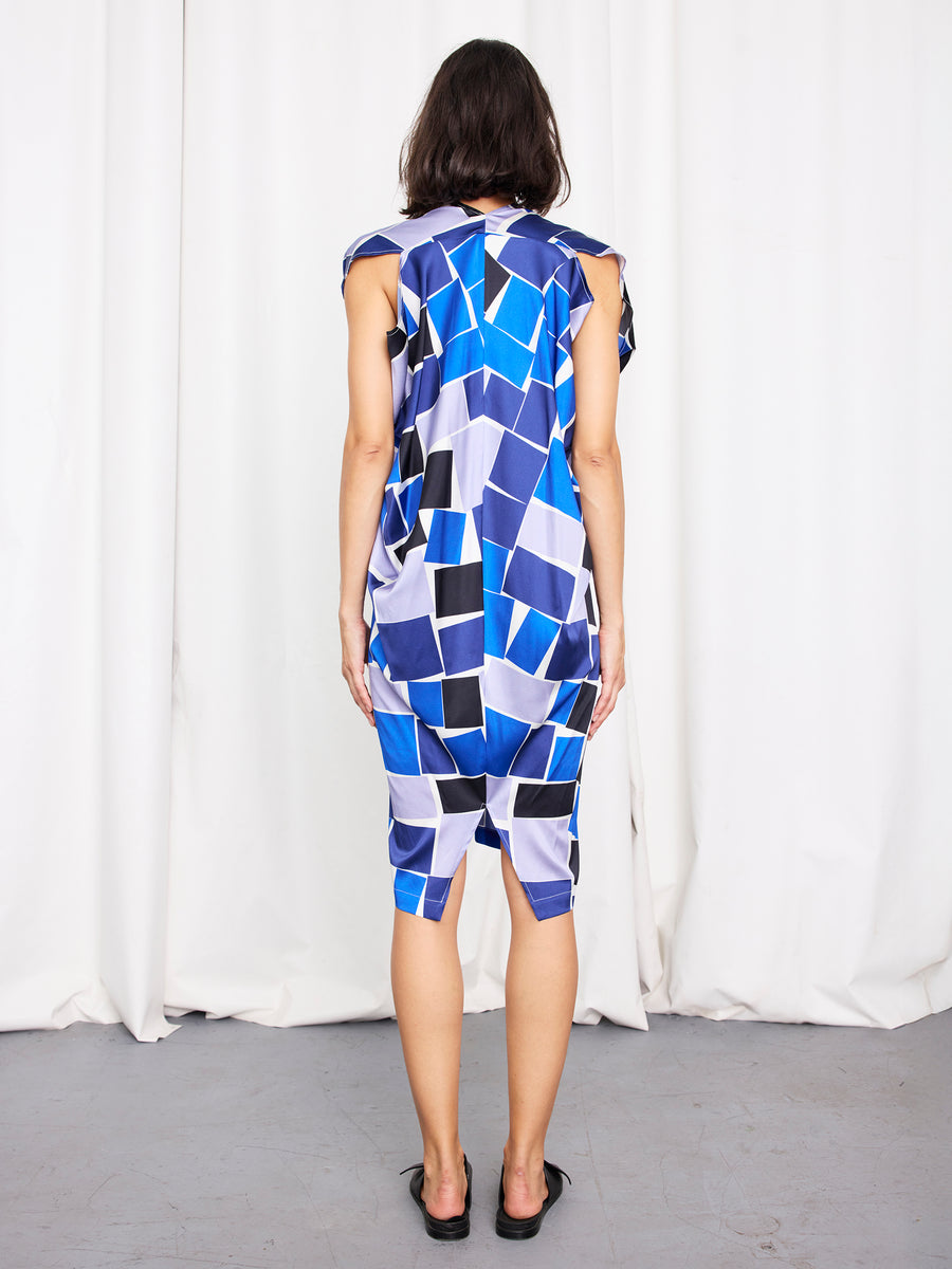 A person faces away, wearing the knee-length Issa Dress by Zero + Maria Cornejo. The stretch silk dress features a blue, black, and white geometric pattern and is paired with black shoes. White curtains hang in the background.