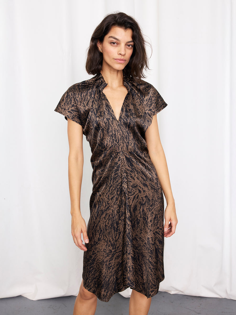 A woman with medium-length dark hair wears the Ruched Stella Dress by Zero + Maria Cornejo, featuring a black and brown abstract print, standing in front of white curtains.