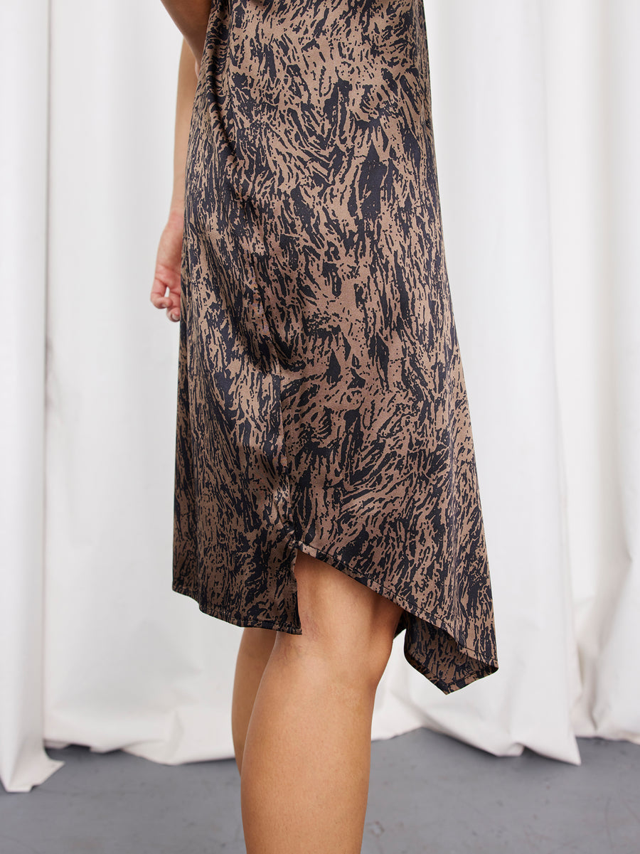 A person stands before white curtains, showing only their lower half, dressed in the Ruched Stella Dress by Zero + Maria Cornejo—a brown and black printed silk piece with an asymmetrical hem.