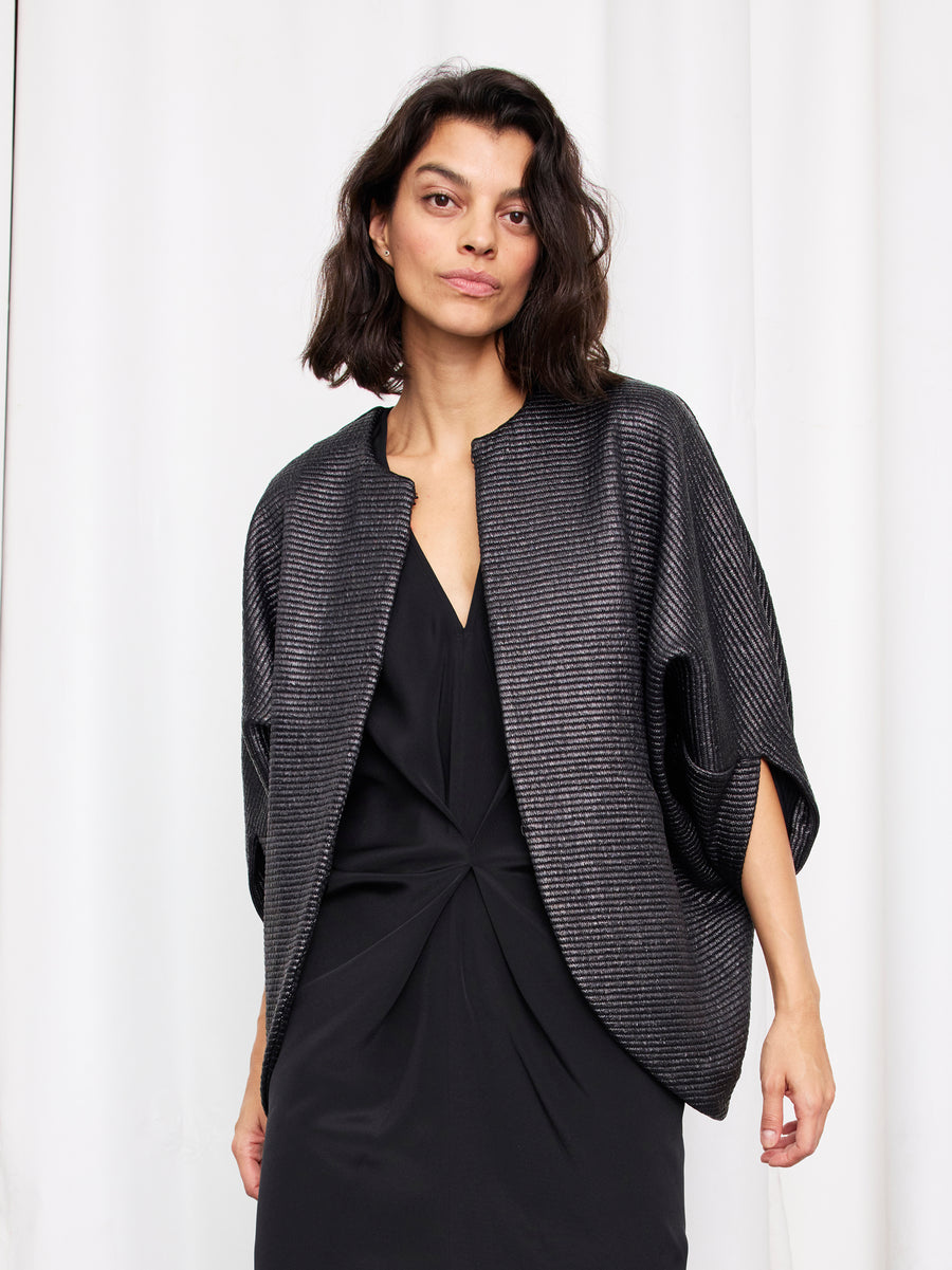 A woman with shoulder-length dark hair wears the Zero + Maria Cornejo Rio Shrug over a knee-length black dress, standing before a white curtain background.