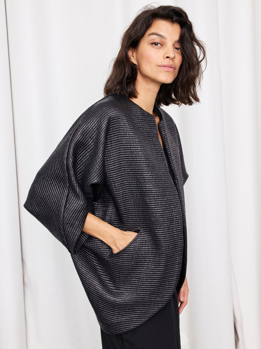 A woman with wavy dark hair wears the Rio Shrug by Zero + Maria Cornejo, a loose, textured woven jacket with pockets and wide sleeves, standing against a white curtain background.