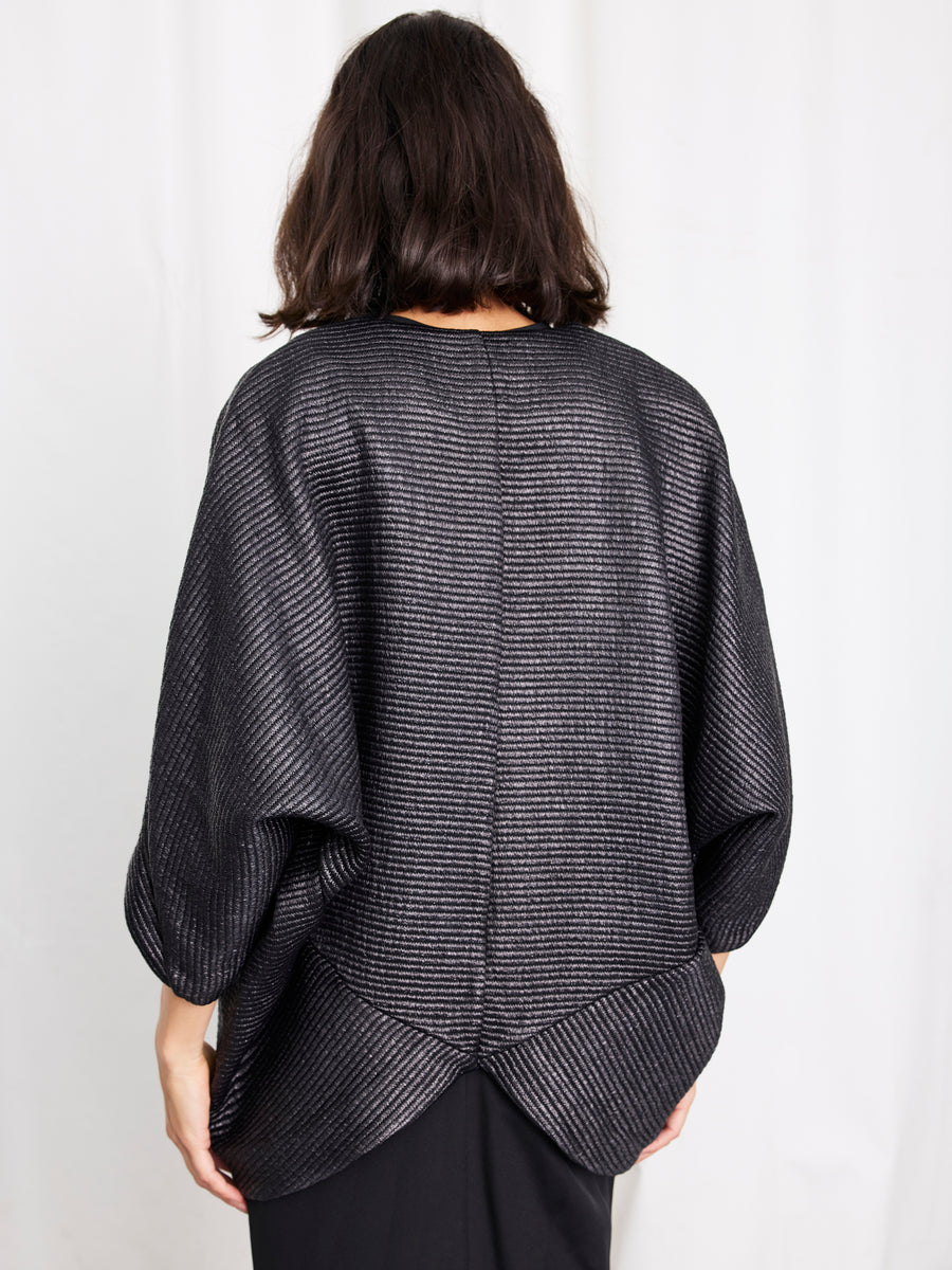 A person with shoulder-length dark hair models the Zero + Maria Cornejo Rio Shrug—a textured, loose-fitting metallic jacket with wide sleeves—standing facing away against a plain white background.