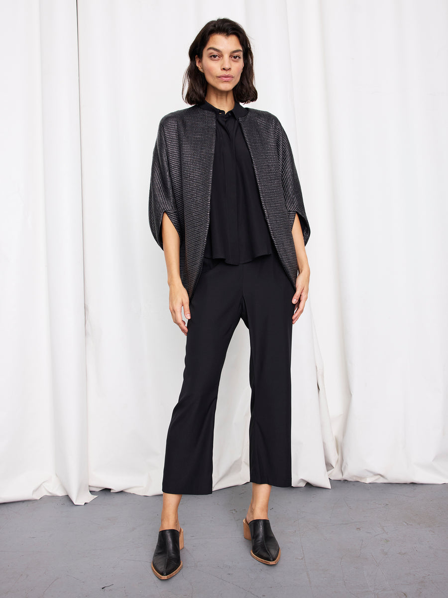 A woman stands before white curtains wearing the metallic Rio Shrug by Zero + Maria Cornejo, paired with a loose black blouse, black pants, and black mules with low wooden heels.