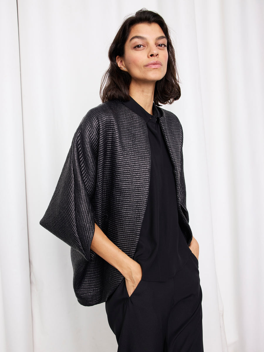 Person with shoulder-length dark hair wears the Zero + Maria Cornejo Rio Shrug over black clothing, standing with hands in pockets against a white curtain background.