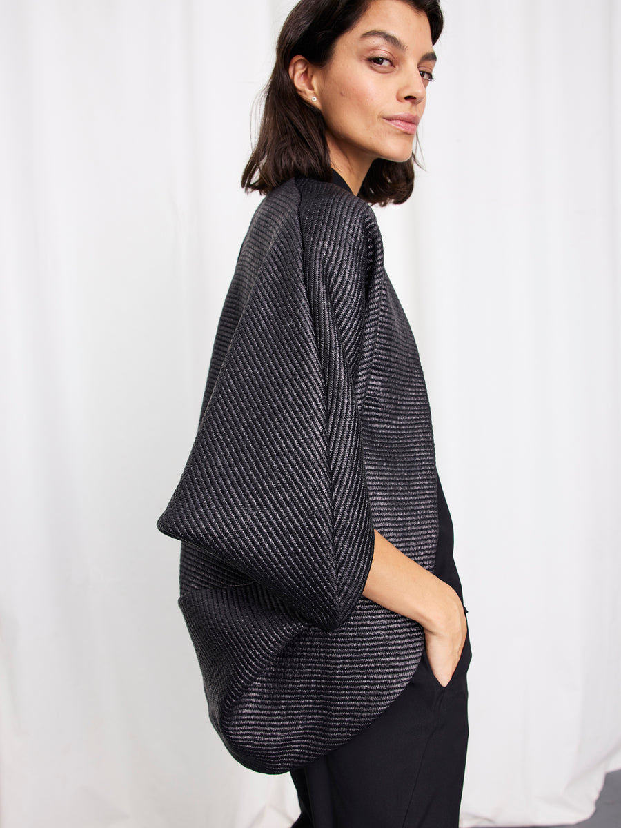 A woman with dark hair wears the Zero + Maria Cornejo Rio Shrug, an oversized metallic draped jacket with wide sleeves, standing sideways with one hand in her pocket against a plain white background.