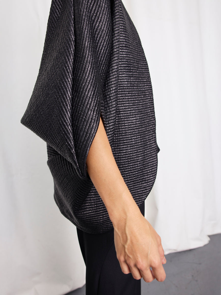 Someone stands against a white curtain, arm relaxed at their side, wearing the Zero + Maria Cornejo Rio Shrug—a textured, loose-fitting black short-sleeve top with diagonal stripes made in New York.