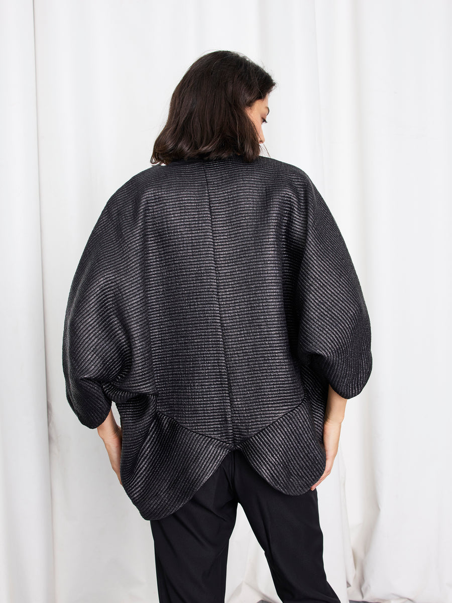 A person with medium-length dark hair stands facing away, wearing the oversized Zero + Maria Cornejo Rio Shrug with rounded sleeves and black pants, in front of white curtains.