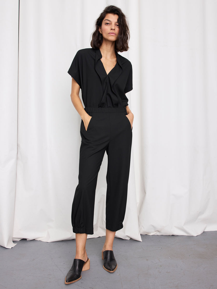 A woman stands in front of white curtains with her hands in her pockets, wearing the Abi Pant by Zero + Maria Cornejo—textured, woven black pants with a cropped fit—paired with black slip-on shoes and a neutral expression.