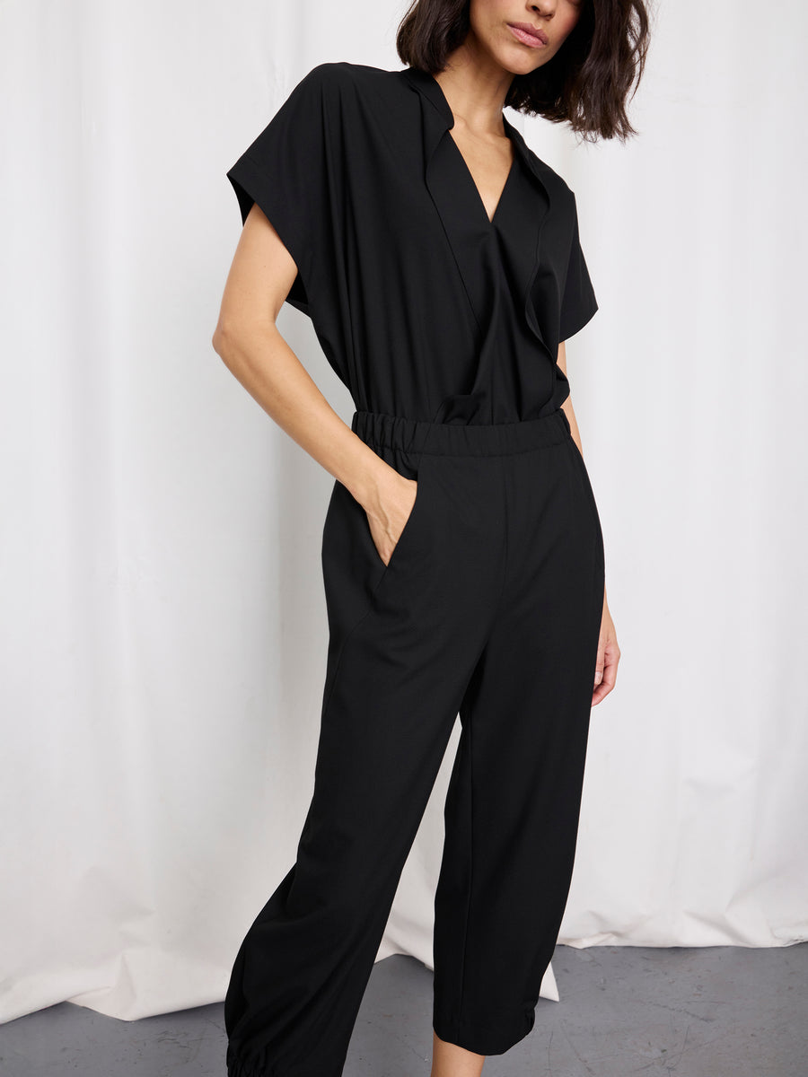 A woman stands against a white backdrop wearing the Zero + Maria Cornejo Abi Pant, featuring textured black fabric, an elastic waistband, cropped legs, and side pockets. Her left hand is in her pocket and her face is partially out of frame.