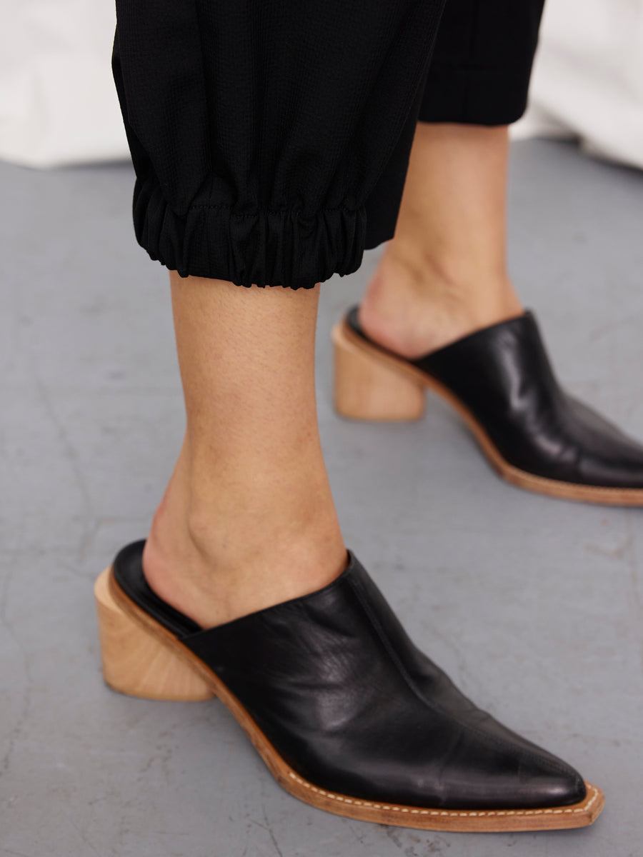 A person wearing the Zero + Maria Cornejo Abi Pant with elastic cuffs and black heeled mules with wooden soles, standing on a gray floor.