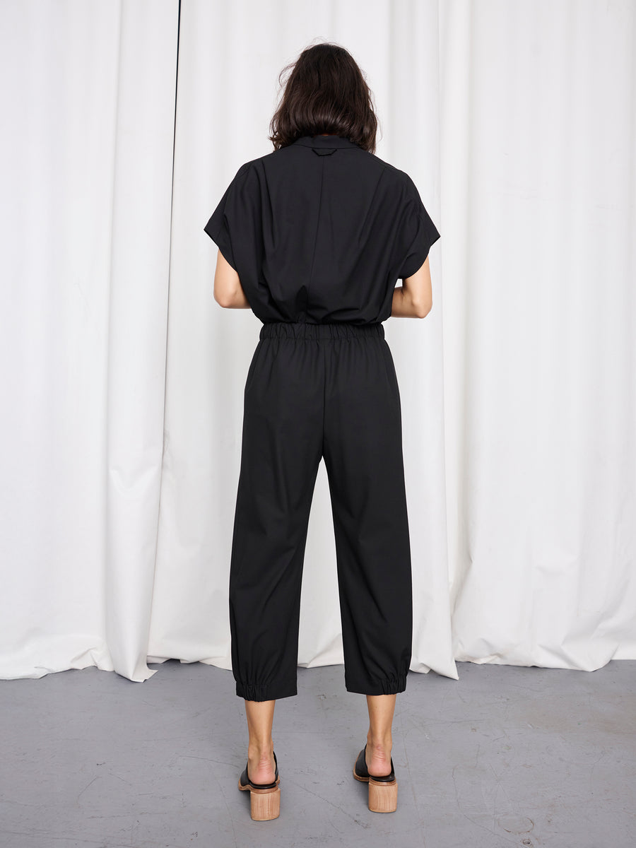 A person with shoulder-length dark hair stands facing away, wearing the Zero + Maria Cornejo Abi Pant, a loose black cropped jumpsuit, paired with wooden-heeled sandals in front of white curtains on a gray floor. Made in New York.