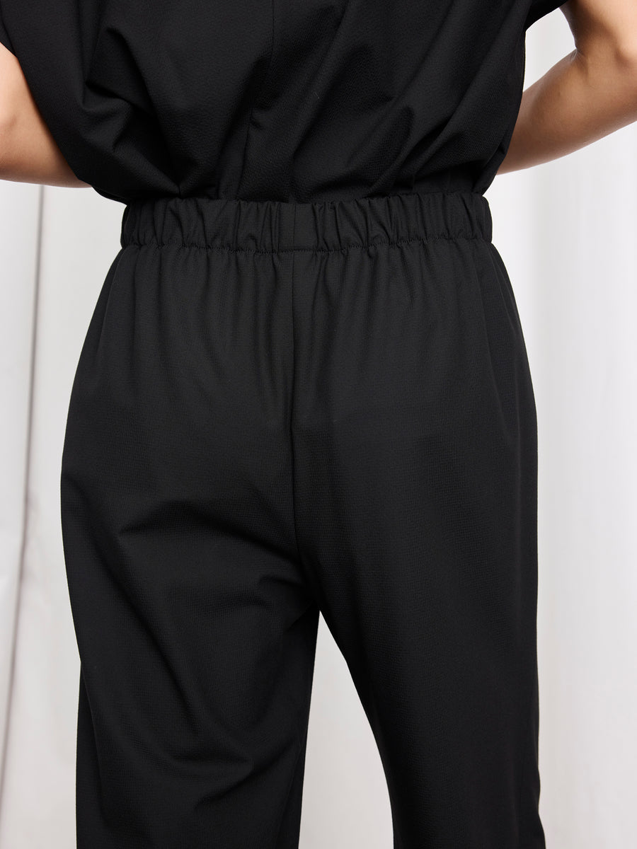 Model wearing the Abi Pant by Zero + Maria Cornejo—loose-fitting, cropped black pants with an elastic waistband, made in New York. Shown from the back and waist down against a plain, light-colored background.