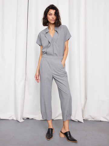 A woman stands in front of white curtains wearing the Abi Pant by Zero + Maria Cornejo, a light gray textured woven cropped pant, styled with black heeled mules and one hand in her pocket, looking relaxed.