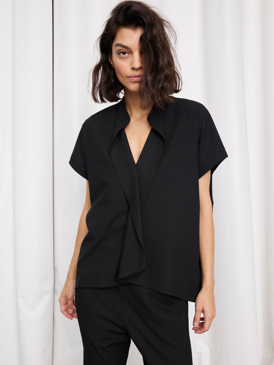 A woman with medium-length dark hair wears the Zero + Maria Cornejo Fin Gaban Shirt, a loose-fitting, short-sleeve black textured woven top, with matching pants, standing before white curtains and looking at the camera neutrally.
