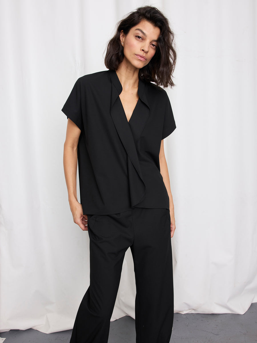 A person with shoulder-length dark hair confidently poses in front of a white curtain, wearing the Zero + Maria Cornejo Fin Gaban Shirt—a loose-fitting, textured woven top with a stand collar—paired with matching black trousers.