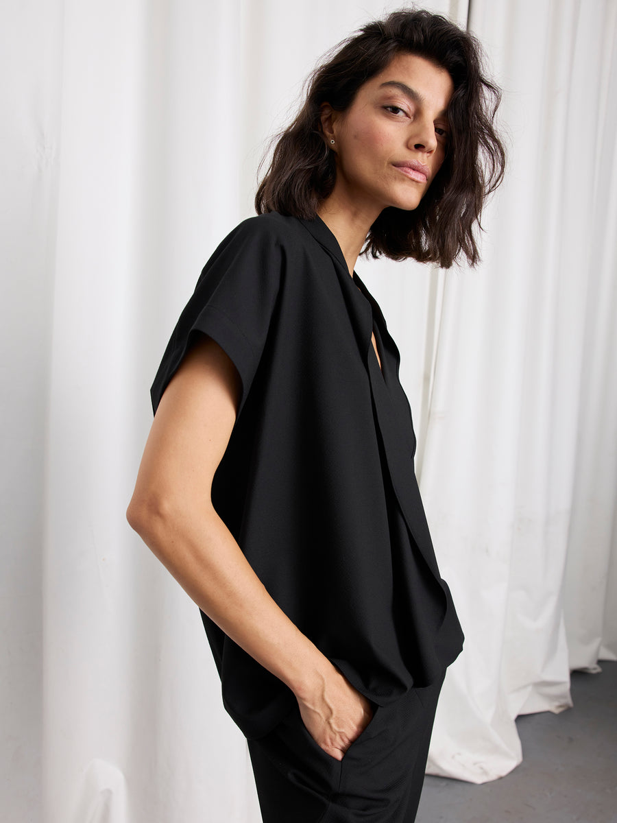 A woman with dark, wavy shoulder-length hair stands sideways in the Fin Gaban Shirt by Zero + Maria Cornejo, posing confidently in front of white curtains—her neutral expression and effortless style reflecting the essence of New York design.