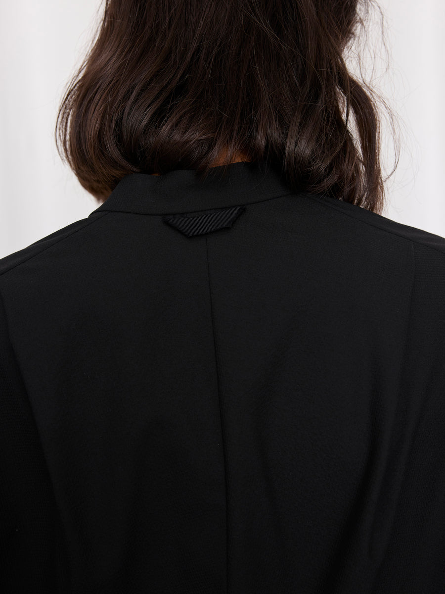 A close-up of a person’s back showcases their dark brown, wavy hair and the upper part of the black, textured Fin Gaban Shirt by Zero + Maria Cornejo, featuring a stand collar against a plain light background.