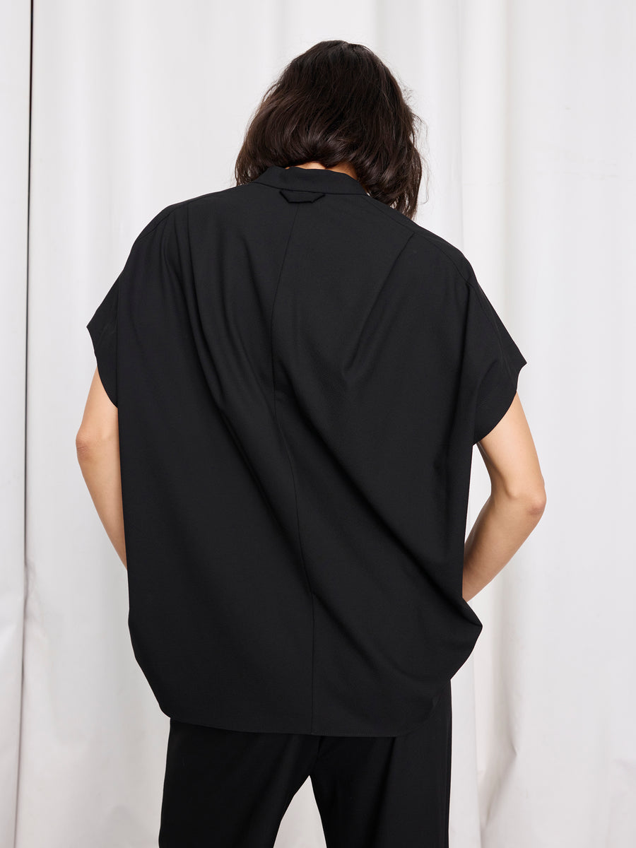 A person with long dark hair stands with their back to the camera, wearing the Zero + Maria Cornejo Fin Gaban Shirt and black pants in front of white curtains.