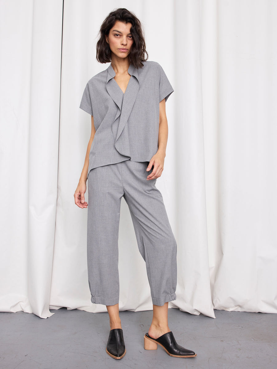 A woman stands before white curtains wearing the Zero + Maria Cornejo Fin Gaban Shirt, a loose-fitting, short-sleeved gray top with an asymmetrical front, paired with matching gray cropped pants and black mule shoes with wooden heels.