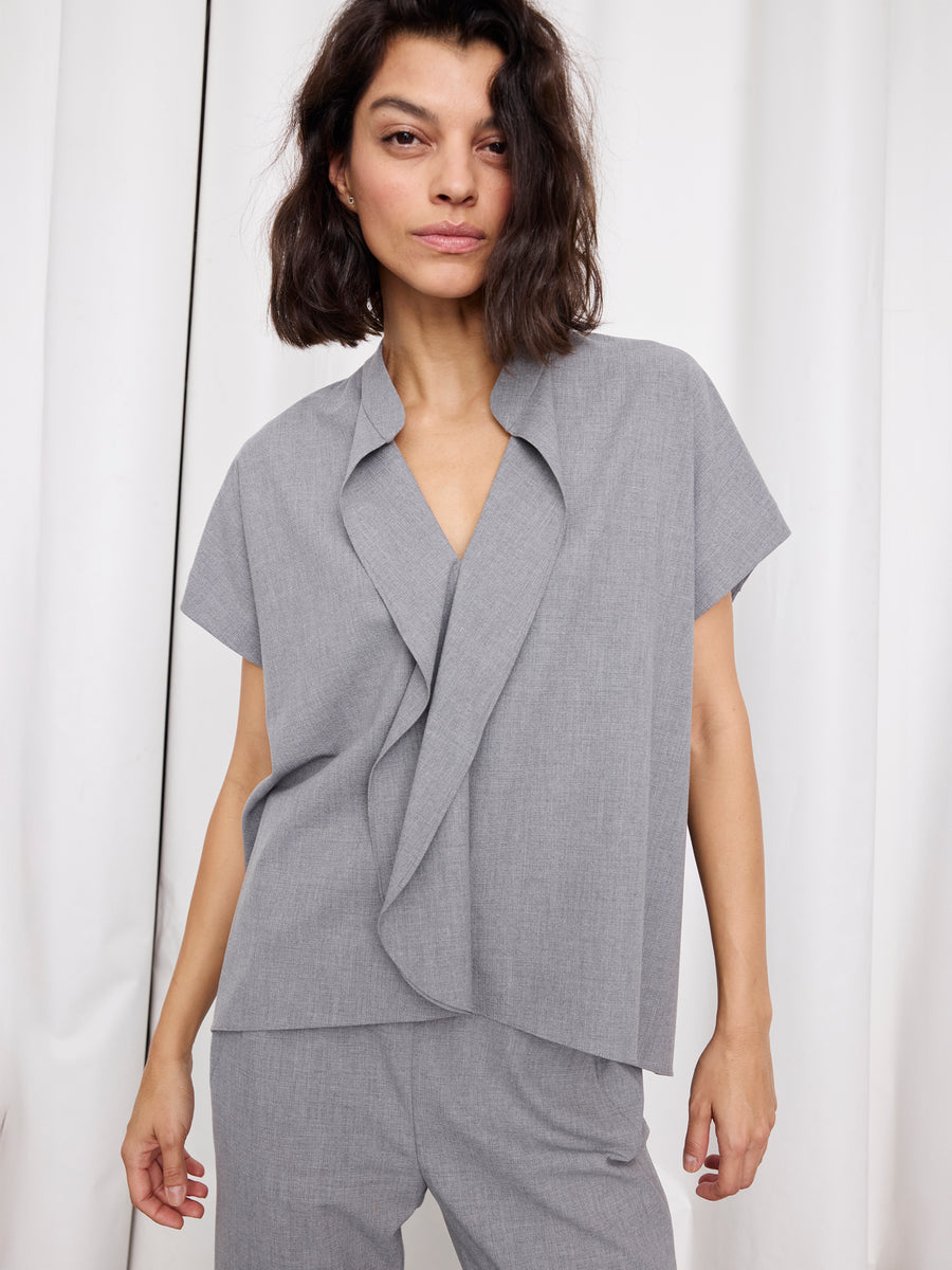 A woman with wavy dark hair wears the Zero + Maria Cornejo Fin Gaban Shirt—a loose, textured woven shirt with a stand collar—paired with matching pants, standing confidently by white curtains. Made in New York.