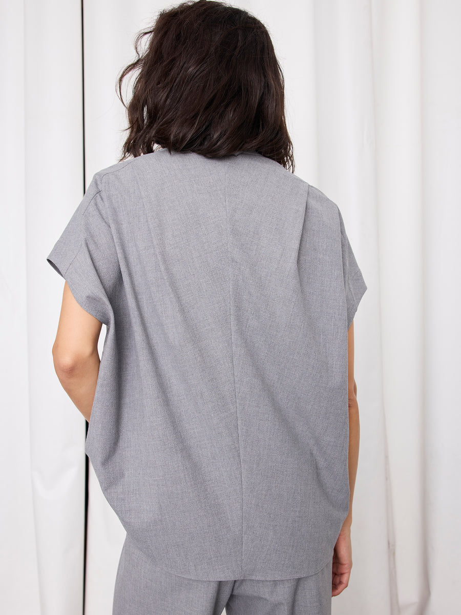 A person with shoulder-length dark hair wears the Fin Gaban Shirt and matching gray pants by Zero + Maria Cornejo, facing away from the camera in front of white curtains, highlighting the shirt’s refined stand collar and New York craftsmanship.
